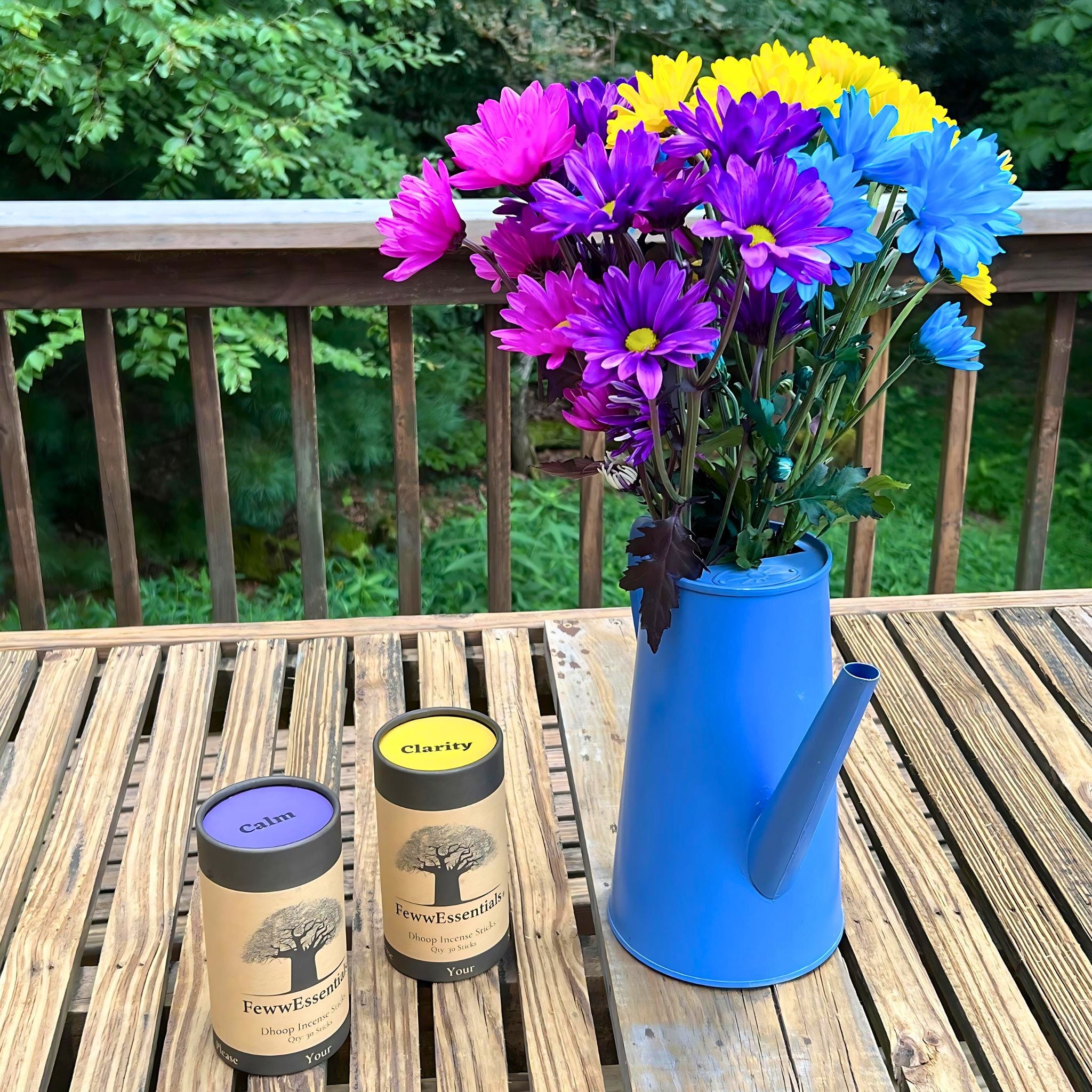 A patio setup with a blue watering can vase holding colorful flowers and FewwEssentials Dhoop Incense Sticks labeled Calm and Clarity on a wooden table.