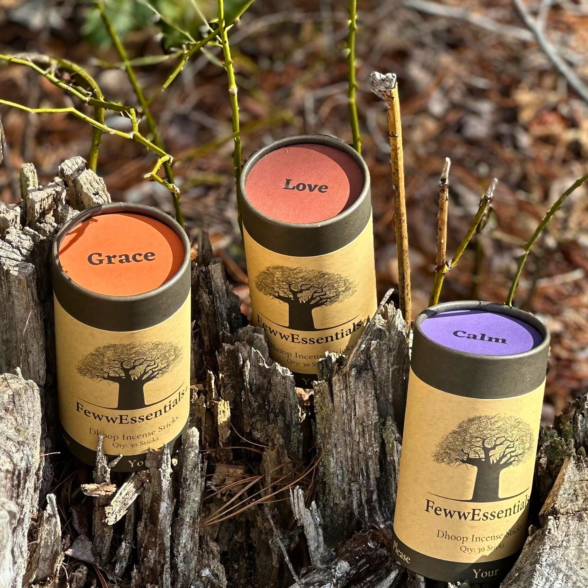 "Close-up of FewwEssentials Dhoop Incense Sticks labeled Grace, Love, and Calm placed on tree stumps with surrounding greenery."