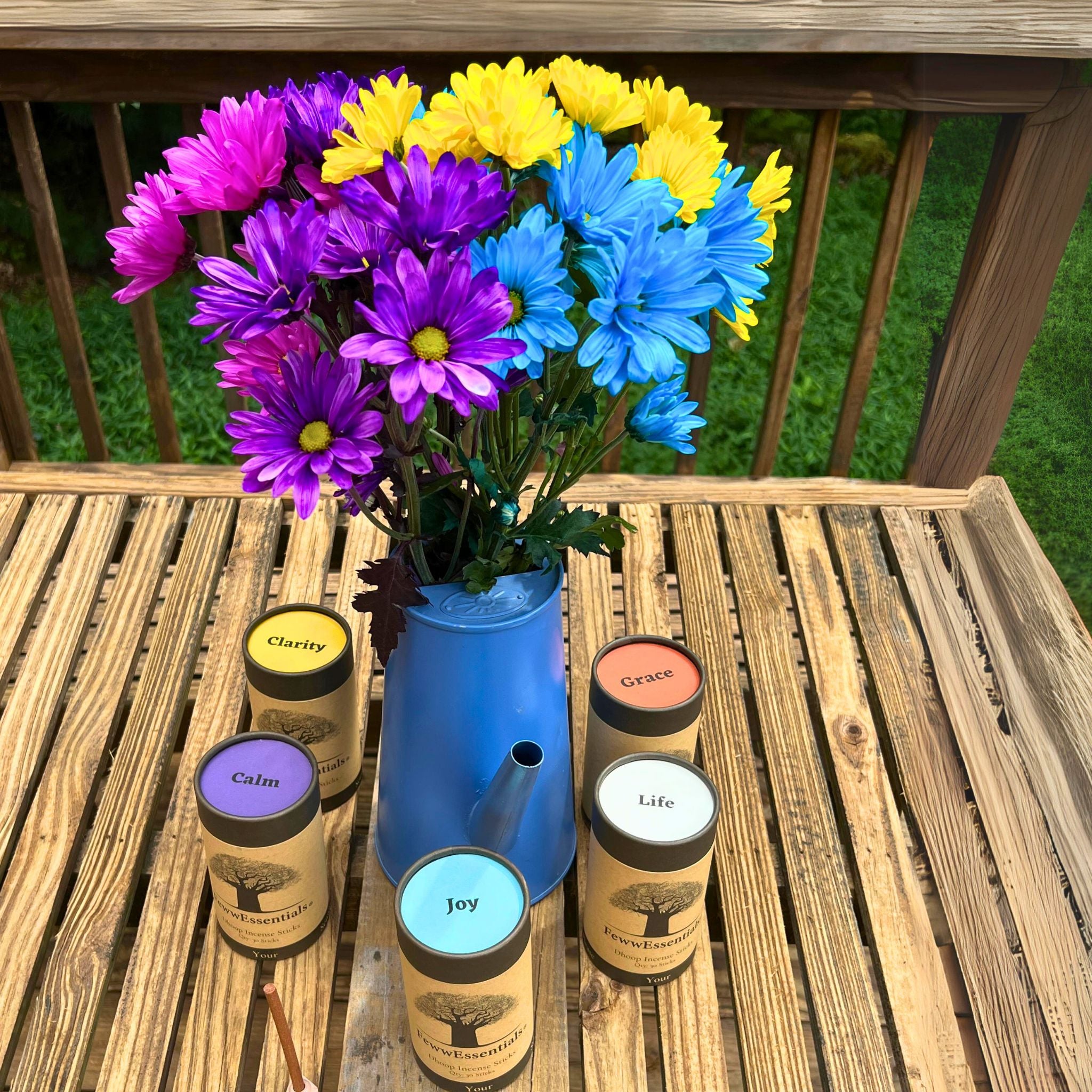 "A wooden table with a blue watering can vase filled with colorful flowers, surrounded by FewwEssentials Dhoop Incense Sticks labeled Clarity, Grace, Calm, Joy, and Life."