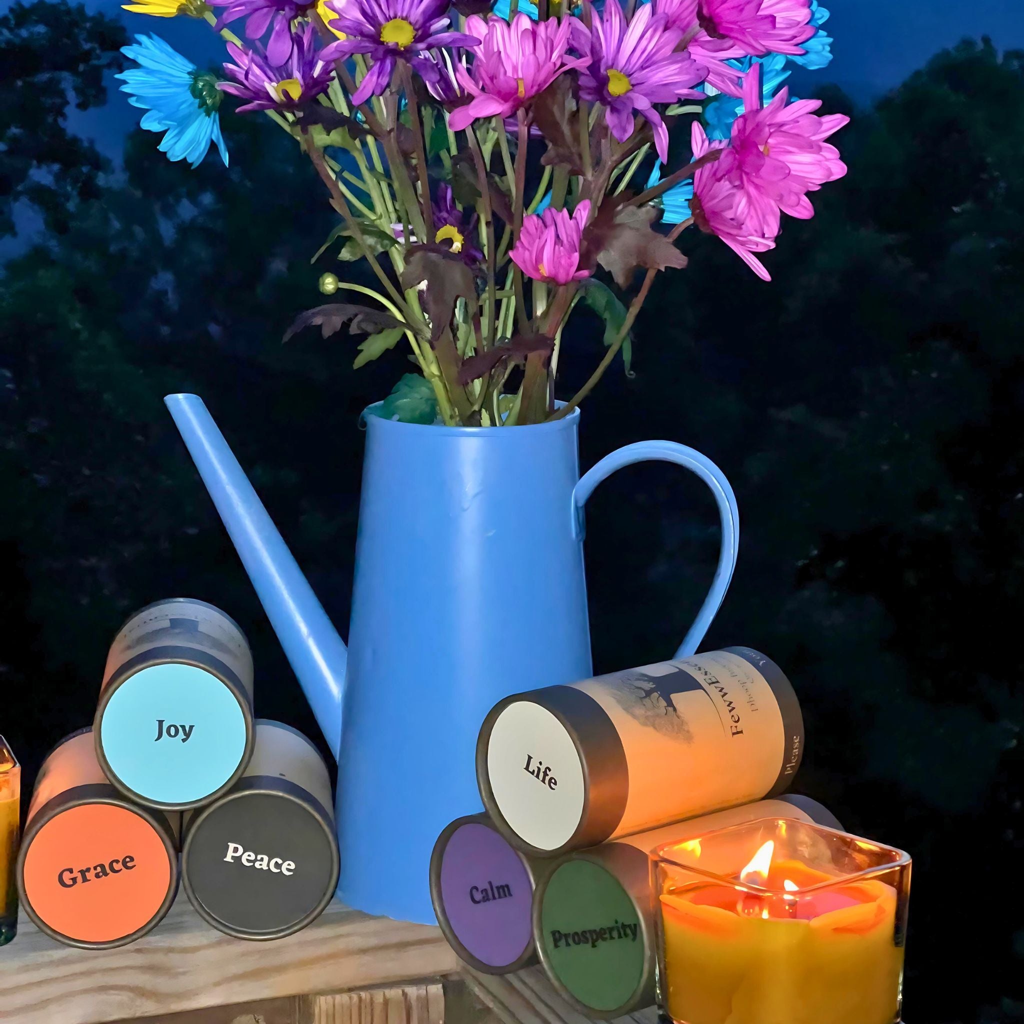 "A vibrant arrangement of FewwEssentials Dhoop Incense Sticks labeled Grace, Joy, Peace, Calm, Prosperity, and Life with a blue watering can vase filled with colorful flowers and a burning candle in a glass holder."