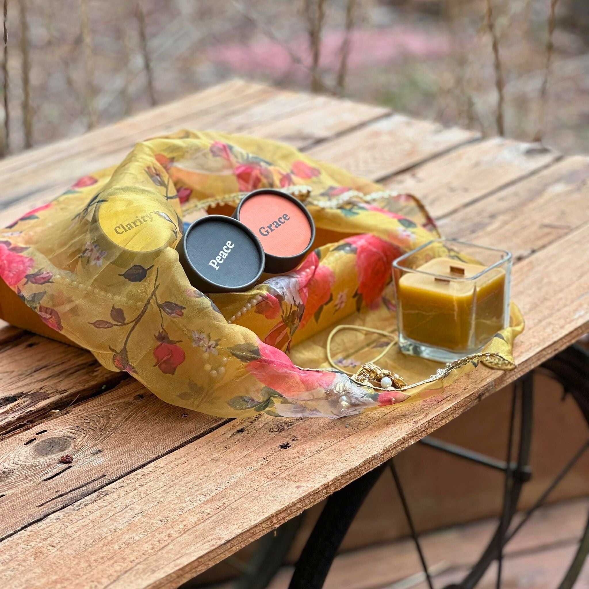 "A rustic outdoor table with a yellow floral scarf, FewwEssentials Dhoop Incense Sticks labeled Clarity, Peace, and Grace, and a square glass candle."